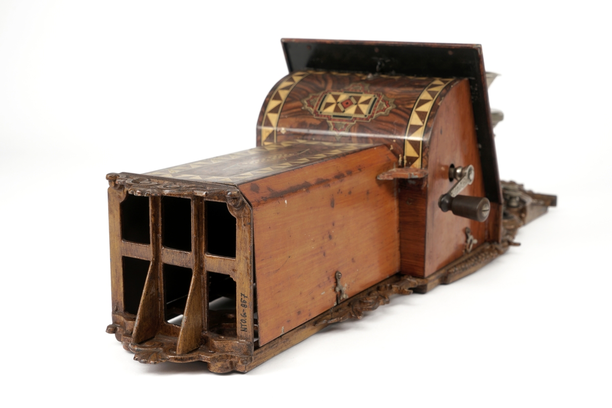 Telefonapparat for kobling på vegg. Fastmontert mikrofon. Høyttaler mangler. Avtagbare, brunlakkerte metalldeksler med dekor. Hengslet topplokk. Smijernsdetaljer. Sveiv med dreid trehåndtak. To bjeller av metall med slagkule mellom disse. Når man dreier på sveiven vil slagkulen "slå" på bjellene og lage en "ringelyd". Diverse koblingspunkter/skruer for tilkobling av ledning.