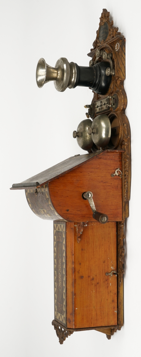 Telefonapparat for kobling på vegg. Fastmontert mikrofon. Høyttaler mangler. Avtagbare, brunlakkerte metalldeksler med dekor. Hengslet topplokk. Smijernsdetaljer. Sveiv med dreid trehåndtak. To bjeller av metall med slagkule mellom disse. Når man dreier på sveiven vil slagkulen "slå" på bjellene og lage en "ringelyd". Diverse koblingspunkter/skruer for tilkobling av ledning.