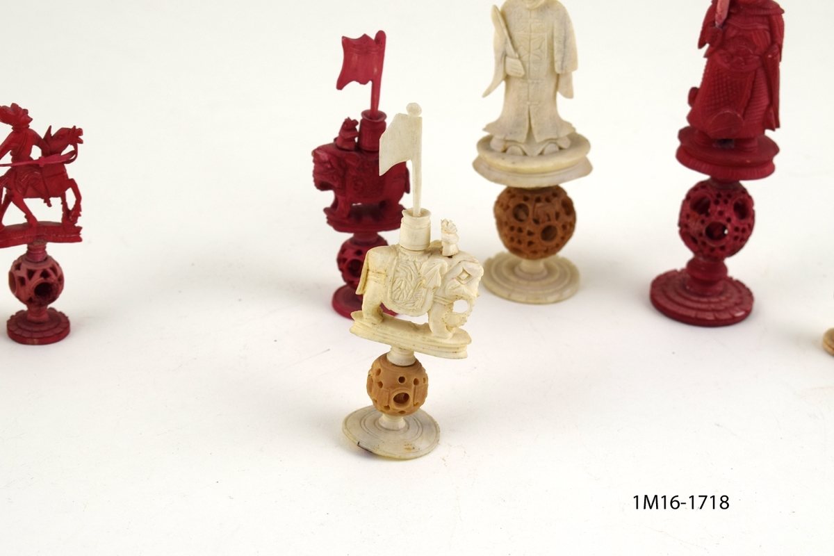 Röda och vita shackpjäser. Totalt 32 figurer. Figurerna i form av människor och elefanter. Minsta figuren 6cm. Största figuren 13 cm.