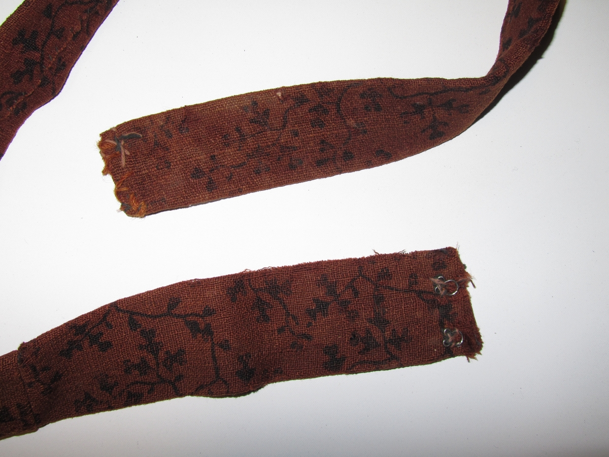 Belte i ull, trykt med svart mønster.  Dobbelt med saum på baksida.  To hekter i kvar ende.  Handsydd. Samansydd av tre delar.