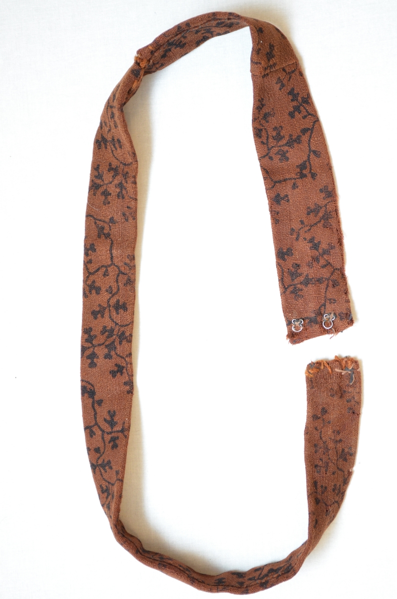 Belte i ull, trykt med svart mønster.  Dobbelt med saum på baksida.  To hekter i kvar ende.  Handsydd. Samansydd av tre delar.