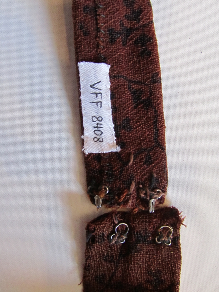 Belte i ull, trykt med svart mønster.  Dobbelt med saum på baksida.  To hekter i kvar ende.  Handsydd. Samansydd av tre delar.