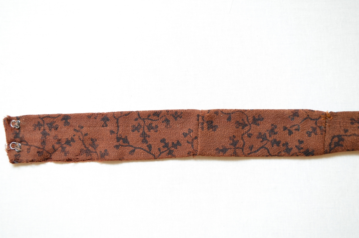 Belte i ull, trykt med svart mønster.  Dobbelt med saum på baksida.  To hekter i kvar ende.  Handsydd. Samansydd av tre delar.