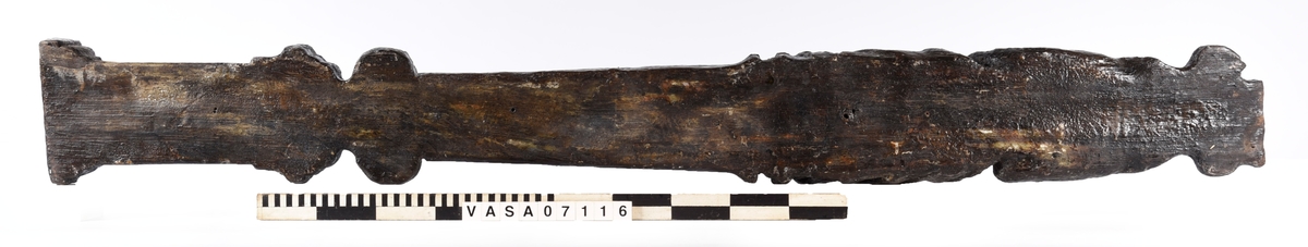 Skulpterad pilaster med övre delen i form av en karyatid med yppig, konstrik frisyr.
Höger arm hålls böjd med handen placerad framför bröstet. Vänster arm hålls utsträckt utefter sidan med handen vilande mot höften. Underkroppen döljs av ett hölje som framtill är prytt med stora, spiralformade ornament och små djurfigurer.
Karyatiden avgränsas från det rullverksornerade pilasterskaftet av en profilerad list. Nedtill avslutas skaftet av ett rullverksornament ovanför den rikt profilerade basen. Nedtill är pilastern försedd med en hög piedestal, prydd med beslagsornamentik. Figuren och dekoren omfattar även pilasterns sidor. Baksidan är slät.


Text in English: Sculpted pilaster with its upper part shaped as a Caryatid with a luxuriant artistic coiffure.
The right arm is held bent with the hand in front of the breast, the left arm is held at the side with the hand resting against the thigh. The lower part of the body is concealed by a shift or slip that is decorated with large spiral-shaped ornaments and small figures of animals.
A profiled batten separates the Caryatid from the shaft of the pilaster, which is ornamented with scrollwork. The shaft terminates with a scrollwork ornament above the richly profiled base. Below, the pilster is equipped with a high pedestal, decked with applied ornamentation. The back is smooth.