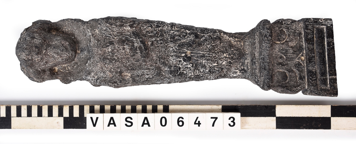 Liten skulpterad pilaster, utformad som en karyatid eller möjligen en putto. Figuren har långt hår och ett knubbigt ansikte, omgivet av en bred krage. Pilasterskaftet är dekorerat med akantusblad. Nedtill avslutas pilastern av en rikt profilerad och dekorerad sockel. Skulpturens baksida är slät.
Skulpturen är mycket sliten.

Text in English: A small carved pilaster, with a caryatid or probably a putto. The figure has a chubby face, long hair and a broad collar. The lower part of the pilaster is decorated with acanthus leaves. The figure stands on a richly profiled and decorated socle. The back of the sculpture is smooth.
The sculpture is very worn.