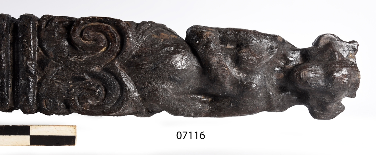 Skulpterad pilaster med övre delen i form av en karyatid med yppig, konstrik frisyr.
Höger arm hålls böjd med handen placerad framför bröstet. Vänster arm hålls utsträckt utefter sidan med handen vilande mot höften. Underkroppen döljs av ett hölje som framtill är prytt med stora, spiralformade ornament och små djurfigurer.
Karyatiden avgränsas från det rullverksornerade pilasterskaftet av en profilerad list. Nedtill avslutas skaftet av ett rullverksornament ovanför den rikt profilerade basen. Nedtill är pilastern försedd med en hög piedestal, prydd med beslagsornamentik. Figuren och dekoren omfattar även pilasterns sidor. Baksidan är slät.


Text in English: Sculpted pilaster with its upper part shaped as a Caryatid with a luxuriant artistic coiffure.
The right arm is held bent with the hand in front of the breast, the left arm is held at the side with the hand resting against the thigh. The lower part of the body is concealed by a shift or slip that is decorated with large spiral-shaped ornaments and small figures of animals.
A profiled batten separates the Caryatid from the shaft of the pilaster, which is ornamented with scrollwork. The shaft terminates with a scrollwork ornament above the richly profiled base. Below, the pilster is equipped with a high pedestal, decked with applied ornamentation. The back is smooth.