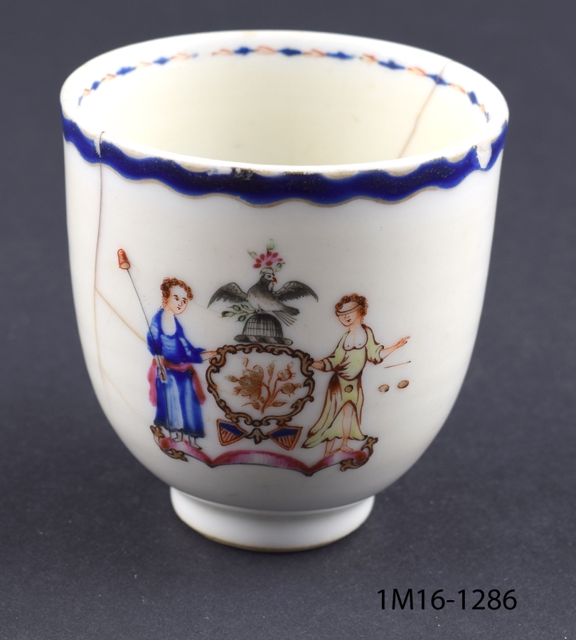 Vit kopp med blå kanter upptill. Motiv mitt på koppen med två kvinnor och en fågel.