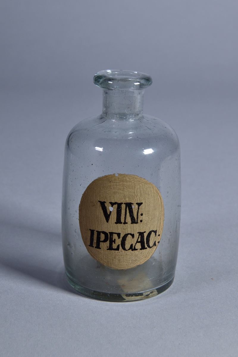 Flaska av klart glas, cylindrisk, kort hals med utsvängd mynning. Påmålad etikett med svart text.