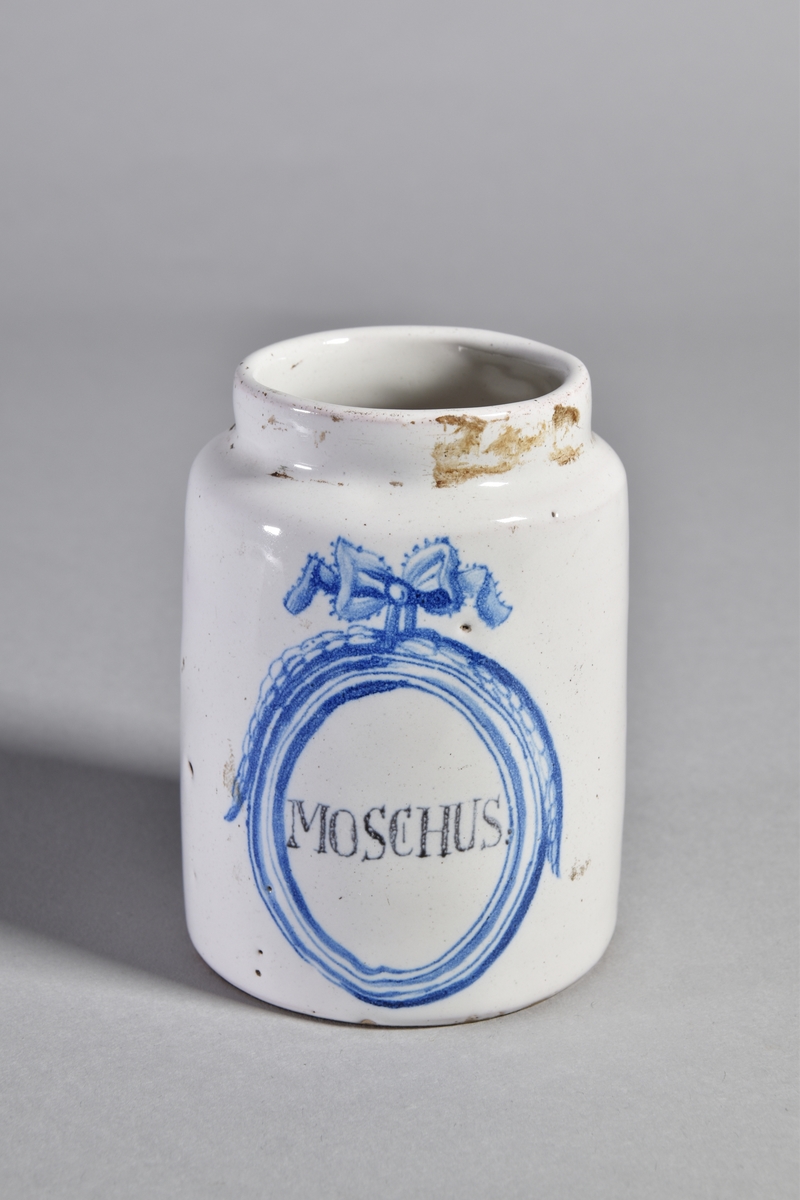 Apoteksburk av vitglaserad fajans, cylindrisk form med indragen skuldra. Svart text i blåmålad medaljong med rosett. Lock av svartmålad bleckplåt, invändig förtenning och pappers skydd.