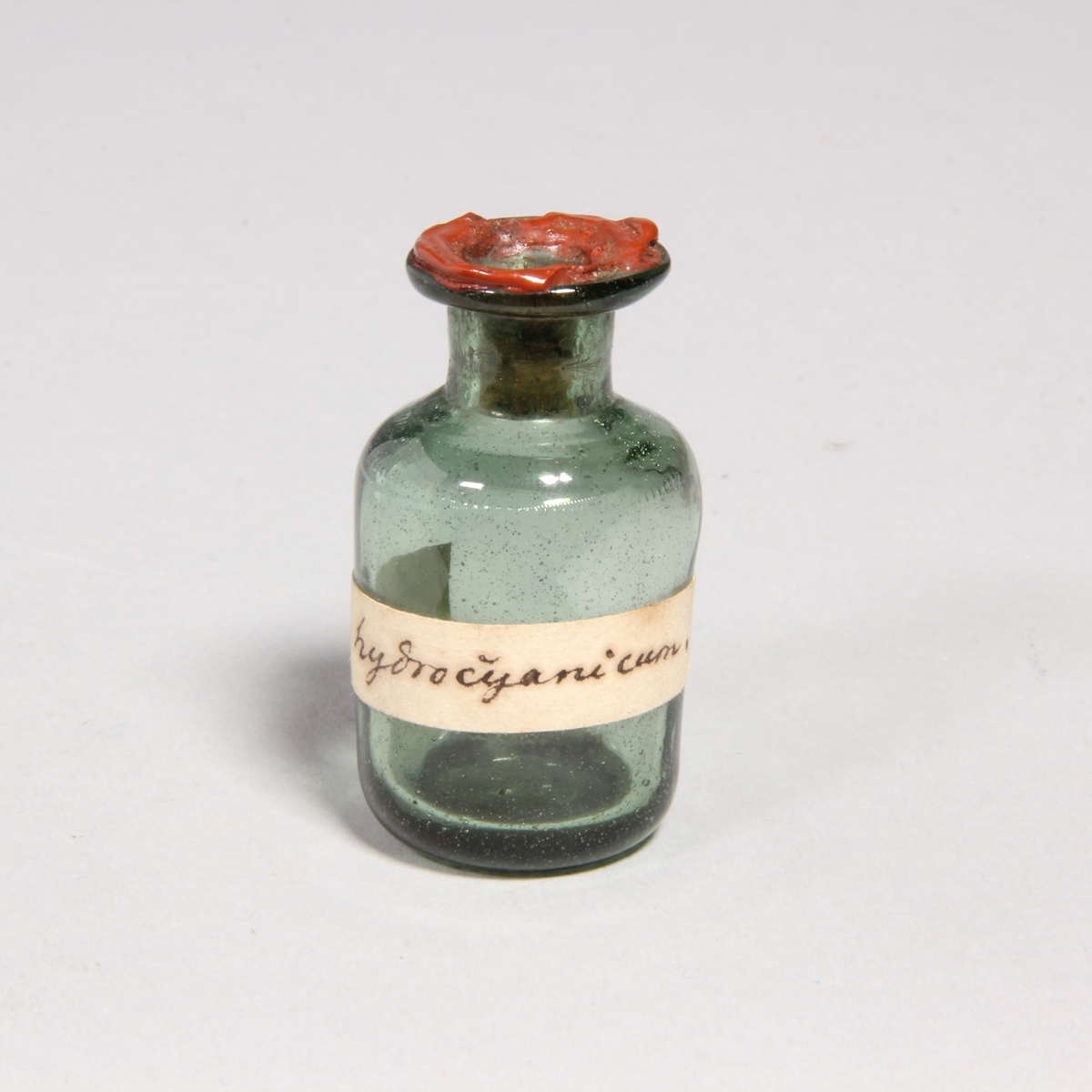 Flaskor, 10 st, av grönt glas med proppar av kork förseglade med rött lack. Står i pappask med lock klädd med marmorerat papper. Flaskorna innehåller rester av cyanid.
