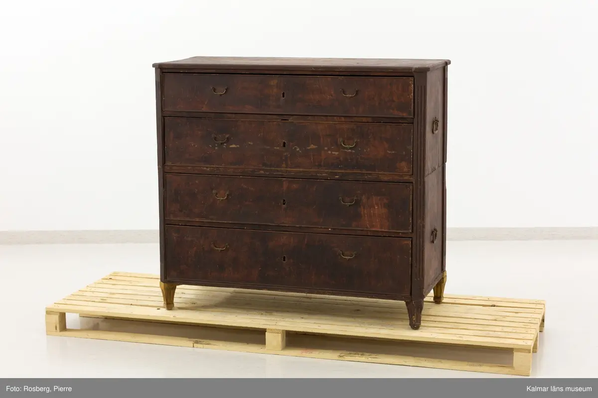 KLM 10747. Kista, dragkista. Av trä, furu, i sengustaviansk stil. Två stapelbara kistor med två lådor vardera istället för lock som på en vanlig kista. Varje del har handtag på kortsidorna så att de går att bära vid flytt. Som möbel fungerar den som en byrå. Räfflade, avskurna hörn och nedåt avsmalnande fötter, varav tre blivit utbytta till nya. Lådornas handtag är av mässing, de på kortsidorna av järn. Möbeln är ådringsmålad i en brun nyans, ej original. Undertill syns ett äldre färglager som går i en grå eller ljusblå nyans.