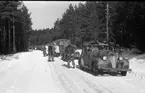 Fordonskolonn. Längst fram en Chevrolet 1939 med punker motorvärmare monterad på vänster sida på fronten. På höger sitter en AGA 104 reflex. Uppe på skärmarna stag till plåtflaggor som brukar nyttjas när regementschefen åker i fordonet. På taket fastsatt i dropplisten ett skidtakräcke gjort i filt och järn för att kunna transportera skidor och stavar. Framför kylaren ett galonliknande material för att skydda från fartvinden som blir extremt kall vid högre hastighet och på så sätt får man en högre temperatur i motorn som ger mer värme till värmepaketet. Med tanke på den fina kamouflagemålningen så är kortet taget från/efter 1941.