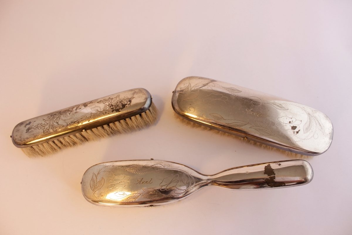 Borstgarnityr bestående av tre borstar en (a) hårborste, två klädesborstar (b-c). Naturborst. Borstarna är klädda i nysilver som har ingraverade stiliserade blommönster. Hårborsten och den ena klädborsten har namnet "Axel" ingraverat på ovansidan. Själva borsthuvudena är tillverkade av trä och sedan har en plåt av nysilver spikats på.