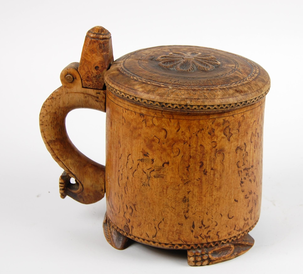 Kanne av dreid valbjørk. Lokk med skjært rosett og to bårder. Tre føtter med utskjæringer.