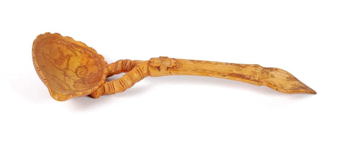 Skje av bjørk. Ovalt skjeblad med åpen tut på den enesiden, profilert langs kanten, kolrosingdekor på oversiden med bl.a. fuglemotiver og blomst, undersiden har kolrosingsdekor av blomster Halsen todelt. Skaftet har kolrosingdekor både på over- og undersiden, ender i en spiss.