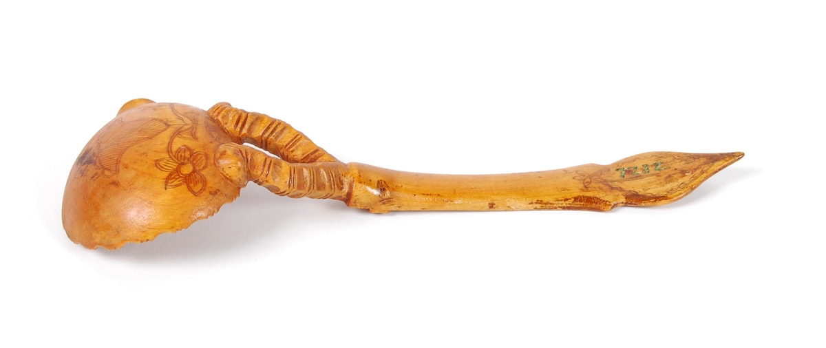 Skje av bjørk. Ovalt skjeblad med åpen tut på den enesiden, profilert langs kanten, kolrosingdekor på oversiden med bl.a. fuglemotiver og blomst, undersiden har kolrosingsdekor av blomster Halsen todelt. Skaftet har kolrosingdekor både på over- og undersiden, ender i en spiss.