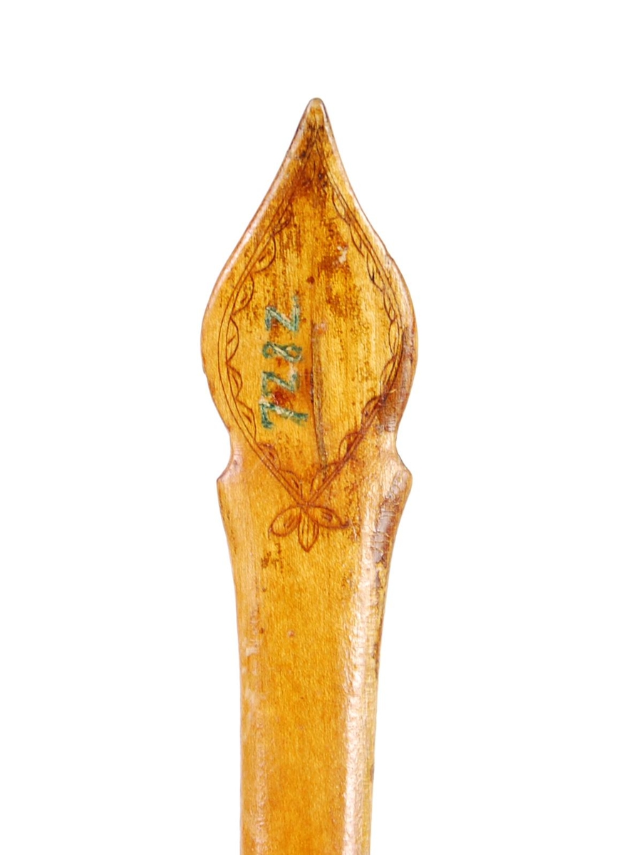 Skje av bjørk. Ovalt skjeblad med åpen tut på den enesiden, profilert langs kanten, kolrosingdekor på oversiden med bl.a. fuglemotiver og blomst, undersiden har kolrosingsdekor av blomster Halsen todelt. Skaftet har kolrosingdekor både på over- og undersiden, ender i en spiss.