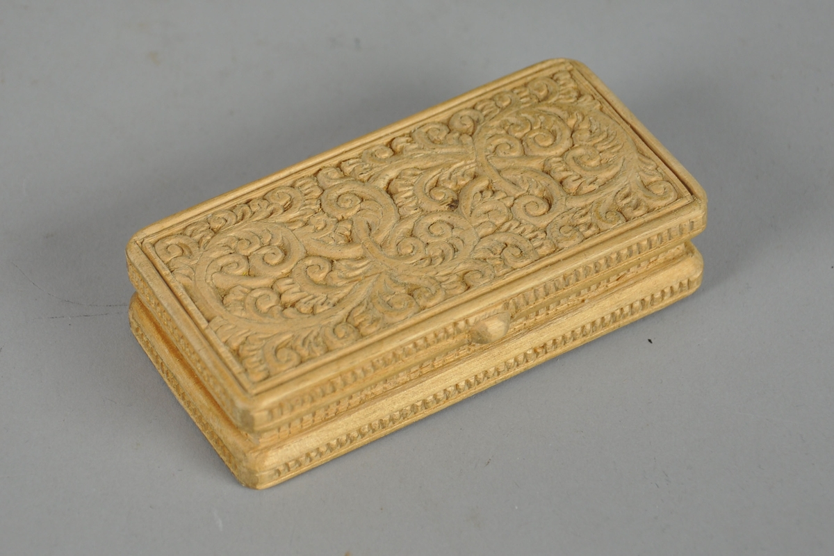 Liten eske av tre, med skjært dekor. Esken har en rektangulær utforming og skjært dekor på lokk og bunn med akantusmotiv, og linjer/rutenett på sidene. 

Esken har en knott på framsiden av lokket.