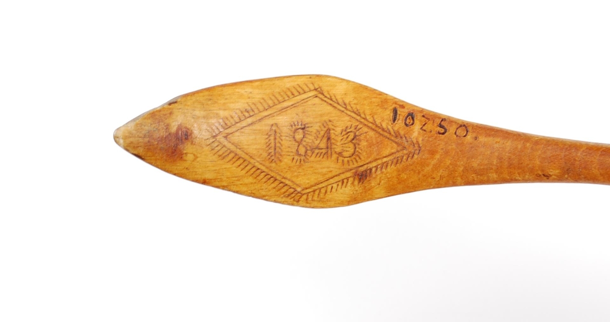 Skje av bjørk. Dypt skjeblad med nebb, i bunnen kolrosingdekor i form av bl.a. en stjerne, undersiden med kolrosingdekor i form av blader og blomster. Buet skaft med kolrosingdekor på over- og undersiden, undersiden har innskrift