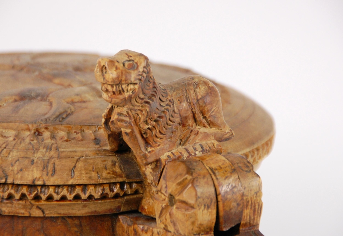 Kanne av dreid valbjørk. Lokket med uskåret springende løve omgitt av bård, bølgete utskjæringer langs kanten. Lokkåpner formet som en løve. Hanken med skjæringer langs kanten, ender i en spiss. Tre liggende løver danner føttene. Buemerke under.