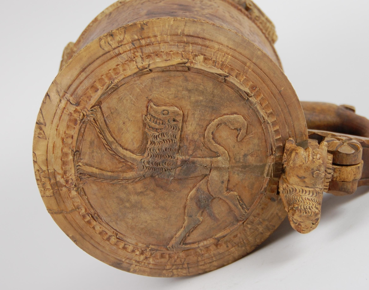 Kanne av dreid valbjørk. Lokket med uskåret springende løve omgitt av bård, bølgete utskjæringer langs kanten. Lokkåpner formet som en løve. Hanken med skjæringer langs kanten, ender i en spiss. Tre liggende løver danner føttene. Buemerke under.