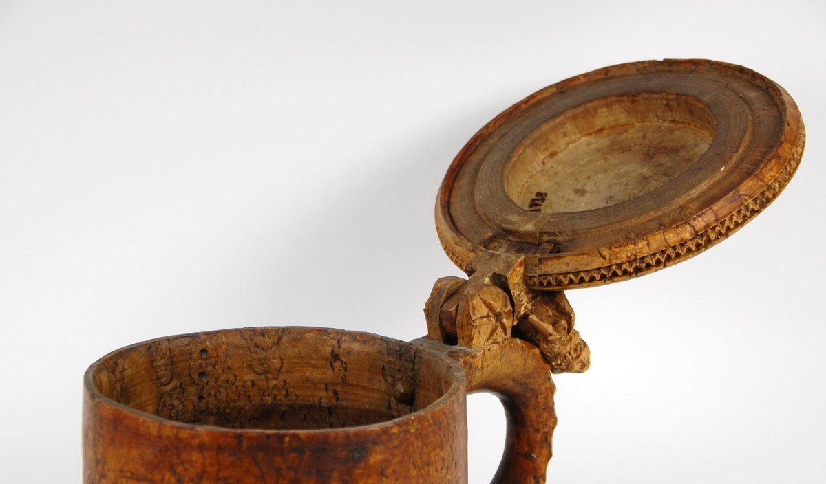 Kanne av dreid valbjørk. Lokket med uskåret springende løve omgitt av bård, bølgete utskjæringer langs kanten. Lokkåpner formet som en løve. Hanken med skjæringer langs kanten, ender i en spiss. Tre liggende løver danner føttene. Buemerke under.