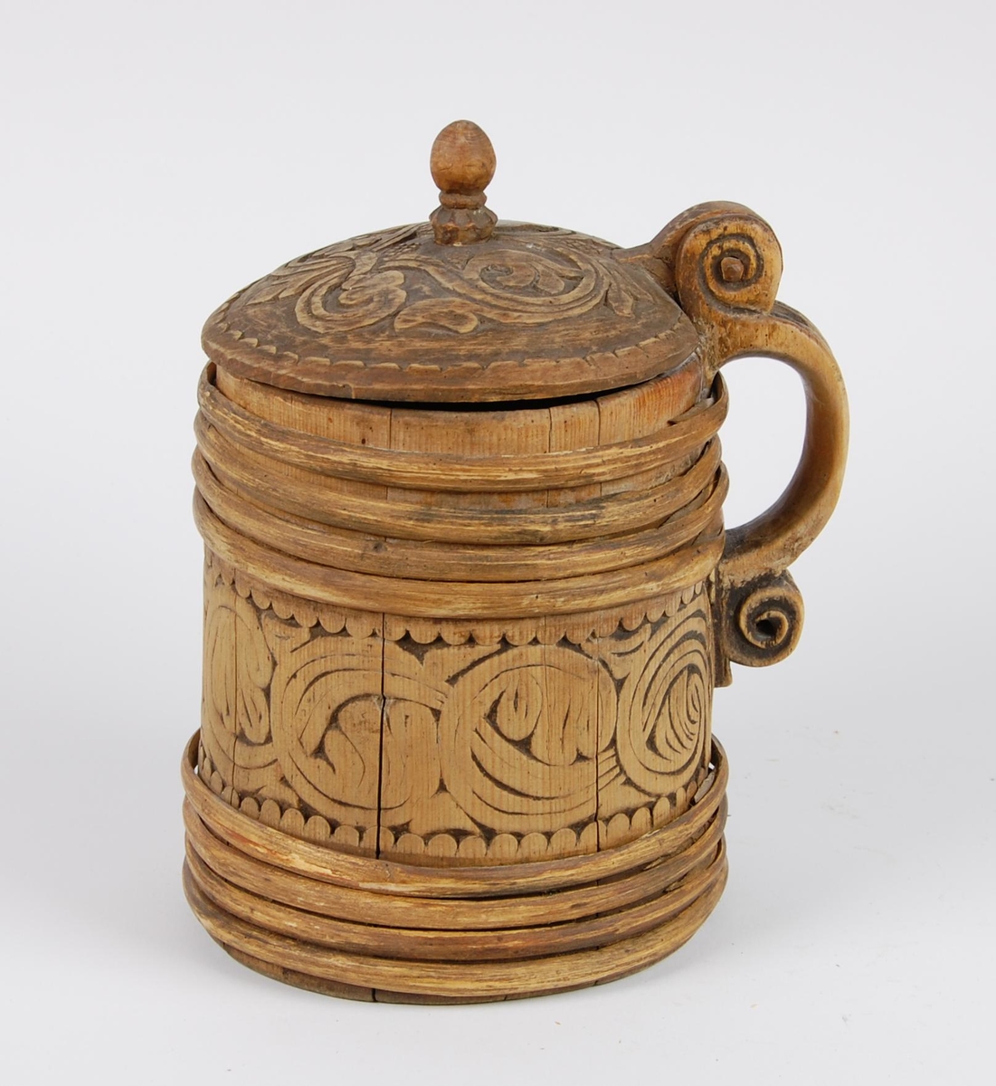 Kanne av trestaver. Lokket med bladlignende ranke, tungeformet bård langs kanten. Lokkgrepet er øverst kuleformet, nederst en ring  med skulpelignende utskjæringer. Hank med spiralformede utskjæringer oppe og nede, er i ett med en av stavene. Korpus har fire gjorder oppe og nede, mellom gjordene en akantusranke, like under og over gjordene tungeformede utskjæringer.