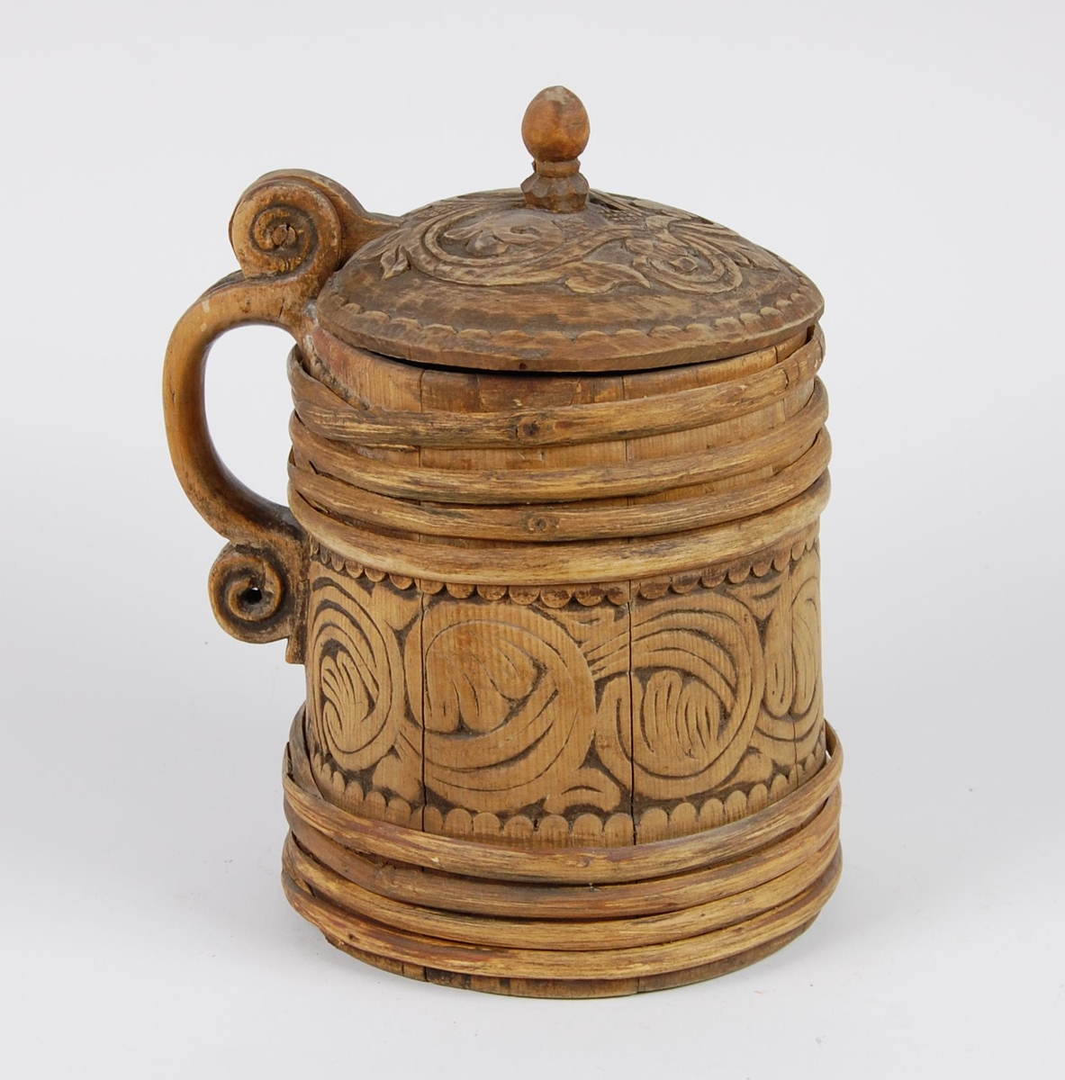 Kanne av trestaver. Lokket med bladlignende ranke, tungeformet bård langs kanten. Lokkgrepet er øverst kuleformet, nederst en ring  med skulpelignende utskjæringer. Hank med spiralformede utskjæringer oppe og nede, er i ett med en av stavene. Korpus har fire gjorder oppe og nede, mellom gjordene en akantusranke, like under og over gjordene tungeformede utskjæringer.