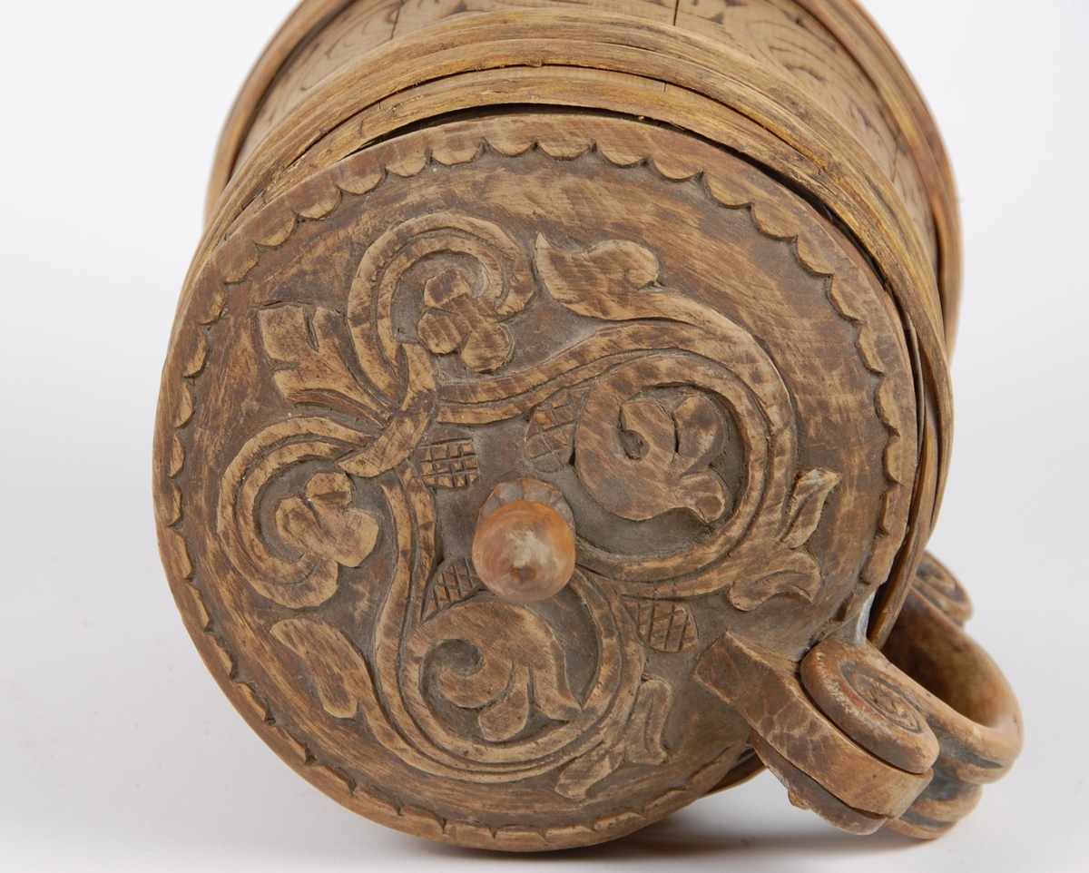 Kanne av trestaver. Lokket med bladlignende ranke, tungeformet bård langs kanten. Lokkgrepet er øverst kuleformet, nederst en ring  med skulpelignende utskjæringer. Hank med spiralformede utskjæringer oppe og nede, er i ett med en av stavene. Korpus har fire gjorder oppe og nede, mellom gjordene en akantusranke, like under og over gjordene tungeformede utskjæringer.