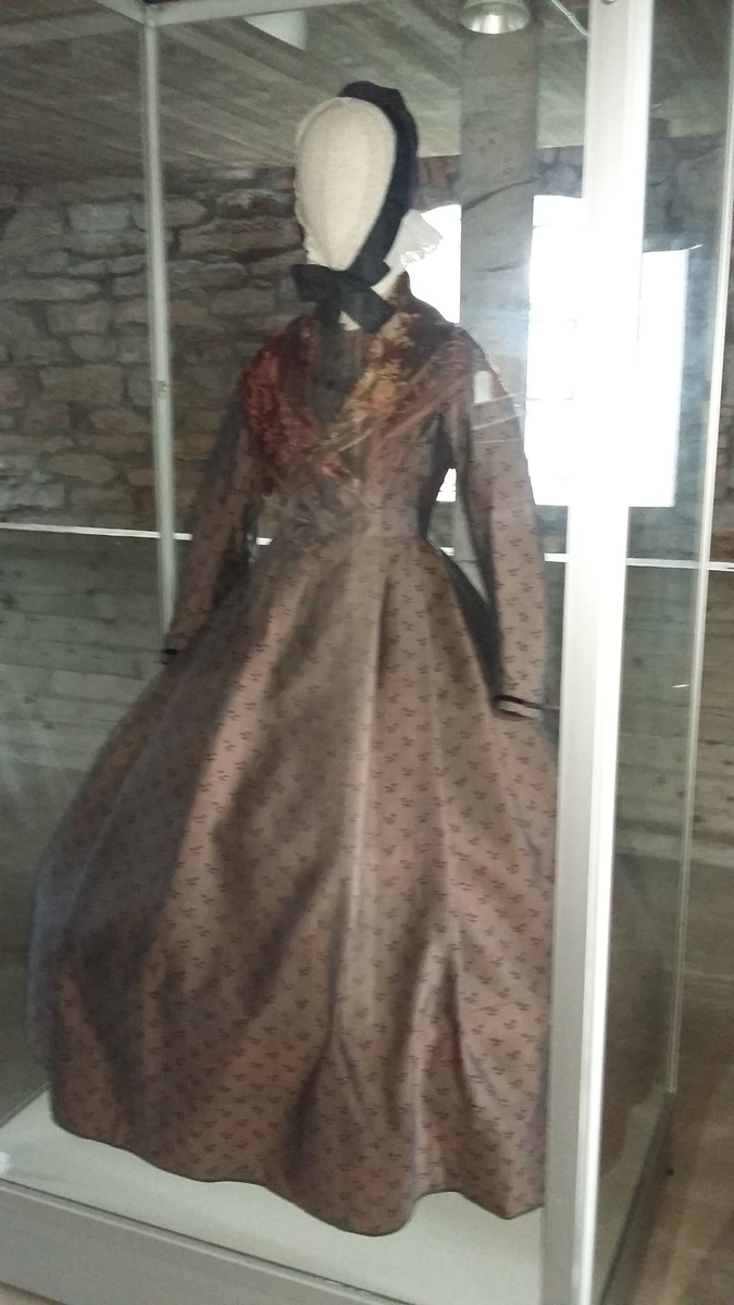 Kjole sammensydd av liv og skjørt med belte i livet med sløyfe i samme stoff. Halvull verkenstoff med trykte blomster. Sydd i Sørum i Akershus ca 1860 og senere brukt på Hamar. Skjørtet har mye håndsøm men vi ser også maskinsøm. Skjørtet er fóret med en skoning av lin i nedre kant. Det er også festet et grønt smalt bånd som slitekant. Overdelen er fóret med ufarget linstoff. Svart fløyelsbånd er festet 1,5 cm fra ermekanten.