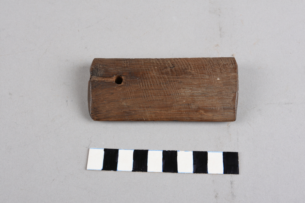 Avlangt trestykke med dråpeforma tverrsnitt, skarp kant på eine langsida og runda på andre.Hol gjennom trestykket, spor i treet frå holet ut til kortenden. Laga i mørkt treverk, ubehandla.