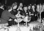 En grupp festklädda kvinnor tar för sig ur en bålskål vid ett elegant dukat bord. Man firar Nordiska kompaniets 50-årsjubileum i januari 1952. Lokalen är sannolikt Stockholms stadshus.