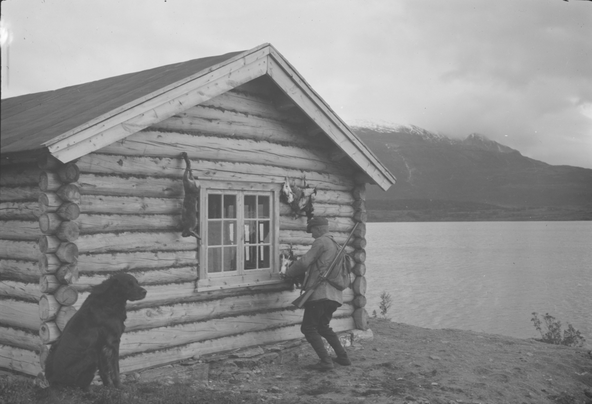 Antatt Sjodalen, jeger med hund og fangst ved hyttevegg.