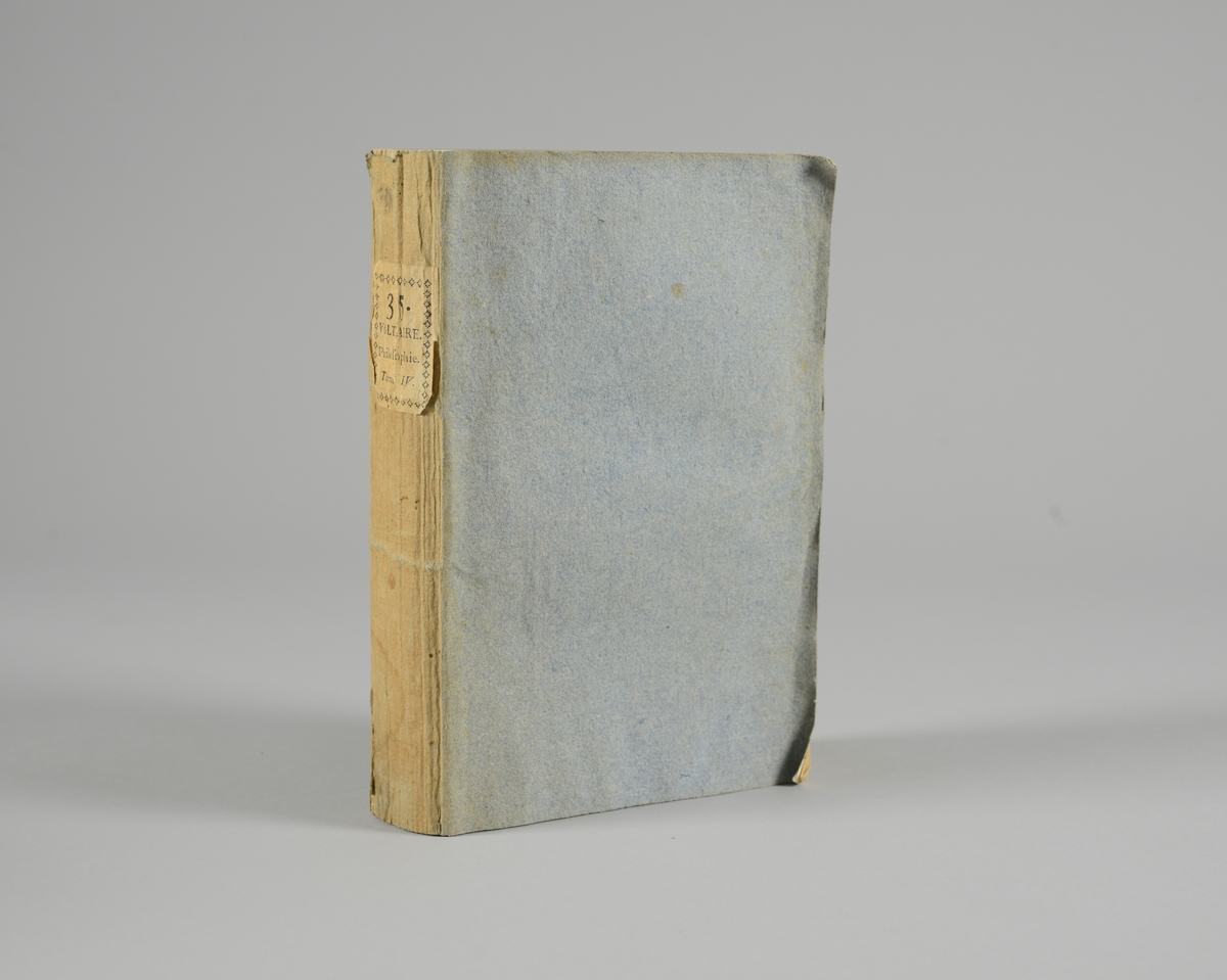 Bok, pappband,"Oeuvres completes de Voltaire", del 35, tryckt1785.
Pärmen klädd med gråblått papper, på pärmarnas insidor klistrade sidor ur annan bok. Med skurna snitt. På ryggen klistrad pappersetikett med tryckt text samt volymens nummer. Ryggen blekt.