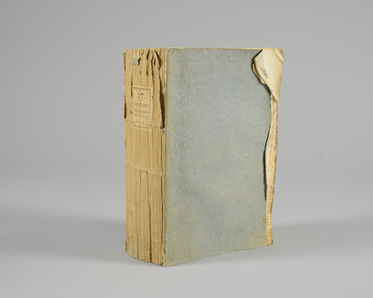 Bok, pappband,"Oeuvres completes de Voltaire", del 37 "Dictionnaire philosophique", tryckt 1785.
Pärmen klädd med gråblått papper, på pärmarnas insidor klistrade sior ur annan bok. Med skurna snitt. På ryggen klistrad pappersetikett med tryckt text samt volymens nummer. Ryggen blekt.