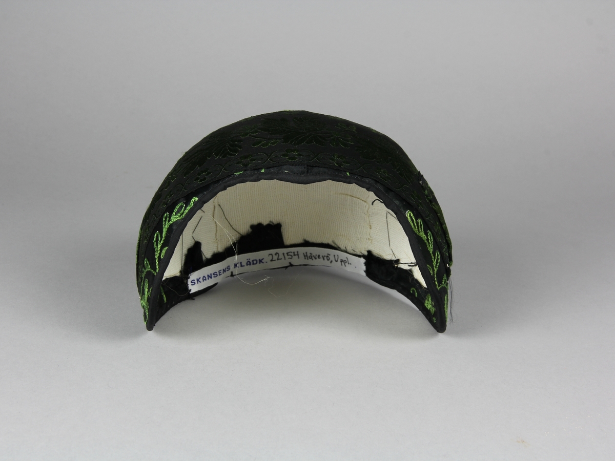 Bindmössa till Häverödräkten. Stomme av vit stärkt tuskaftsvävt tyg som sytts ihop av tre delar och pressats. Klätt med mörkgrönt, nästan svart, sidentyg dekorerat med fantasifulla blommor och kvistar broderade med grön sidentråd i tambursöm. Runt hela mössan är ett 5 cm brett siden band fastnålat. Bandet är svart med grönt mönster. Mössans kanter är klädda med ett tunt svart sidenband som sytts fast längs kanten.