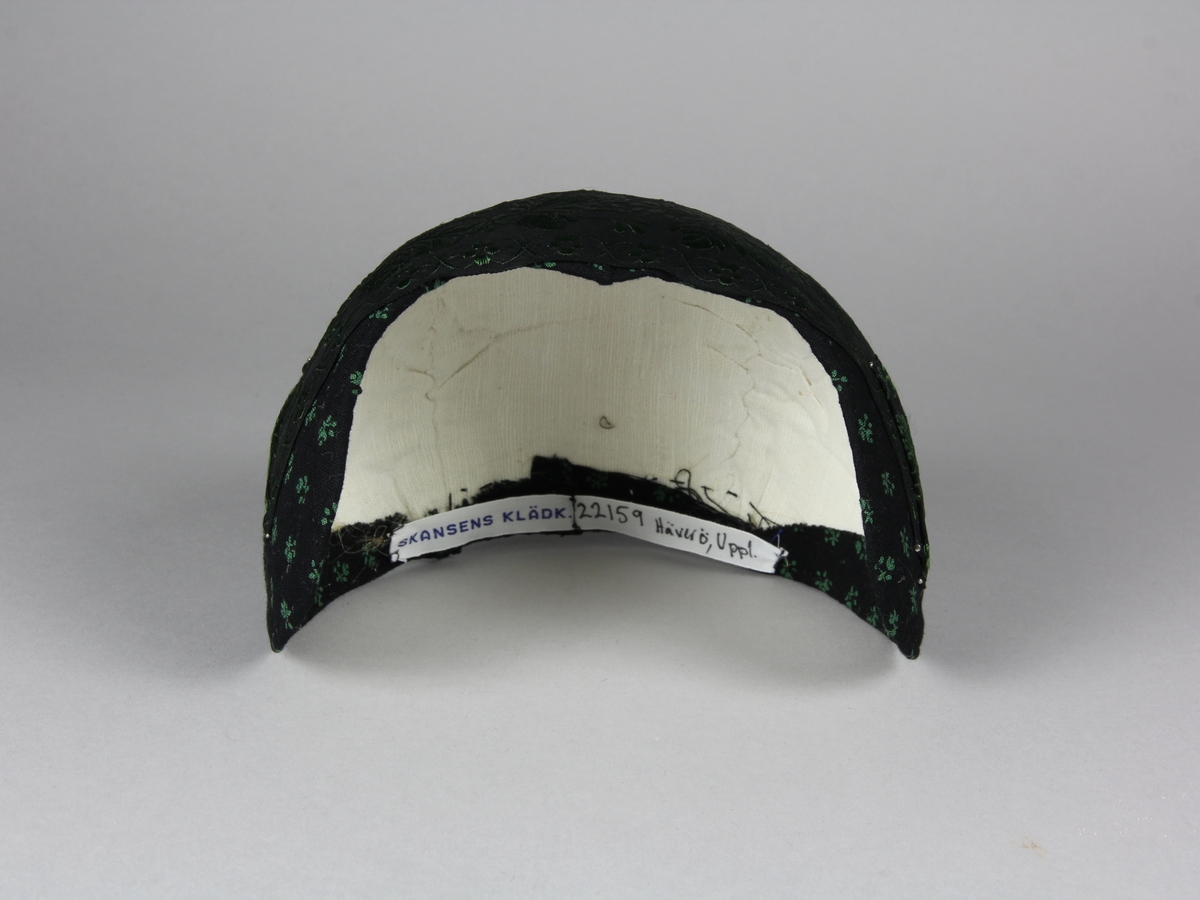 Bindmössa till Häverödräkten. Stomme av vit stärkt tuskaftsvävt tyg som sytts ihop av tre delar och pressats. Klätt med svart bomullstyg med tryckt grönt mönster av små blommor. Runt hela mössan är ett 5 cm brett sidenband fastnålat. Bandet är svart med grönt mönster. Mössans kanter är stickade med prickstygn i två rader.
