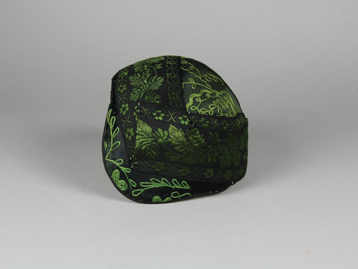Bindmössa till Häverödräkten. Stomme av vit stärkt tuskaftsvävt tyg som sytts ihop av tre delar och pressats. Klätt med mörkgrönt, nästan svart, sidentyg dekorerat med fantasifulla blommor och kvistar broderade med grön sidentråd i tambursöm. Runt hela mössan är ett 5 cm brett siden band fastnålat. Bandet är svart med grönt mönster. Mössans kanter är klädda med ett tunt svart sidenband som sytts fast längs kanten.