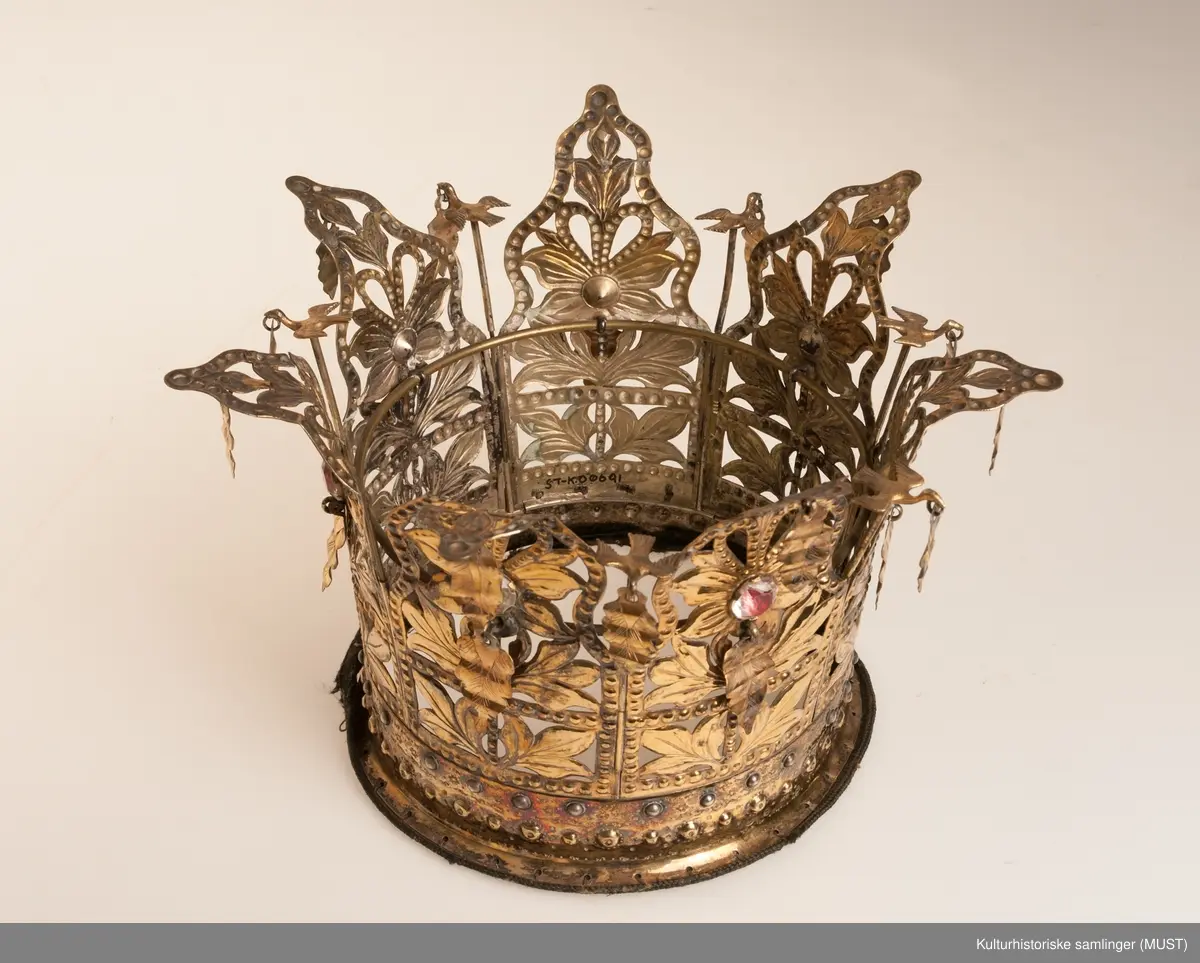 Brudekrone i forgylt sølv. Presset arbeide. Forholdsvis bred bærering med to rader små bukler og ornamentlinjer slått inn med med blad - og blomstformede punsler. Til bæreringens overkant er fastklinket sju gjennombrutte takkestykker som er sammenhengslet. Mønsteret i hvert takkestykke består av en blomst med blader og en foliert glassten. Over blomsten et hjerte, dessuten er til hver takke festet to riflede løv. Mellom takkene står en fugl med utbredte vinger og et løv i nebbet. Stemplet på utsiden av bæreringen med Bergens bymerke og Hoffs guardeinmerke ( stempelmerke fra offentlig ansatt mann som på forlangende skulle prøve finheten av edle metaller ). 