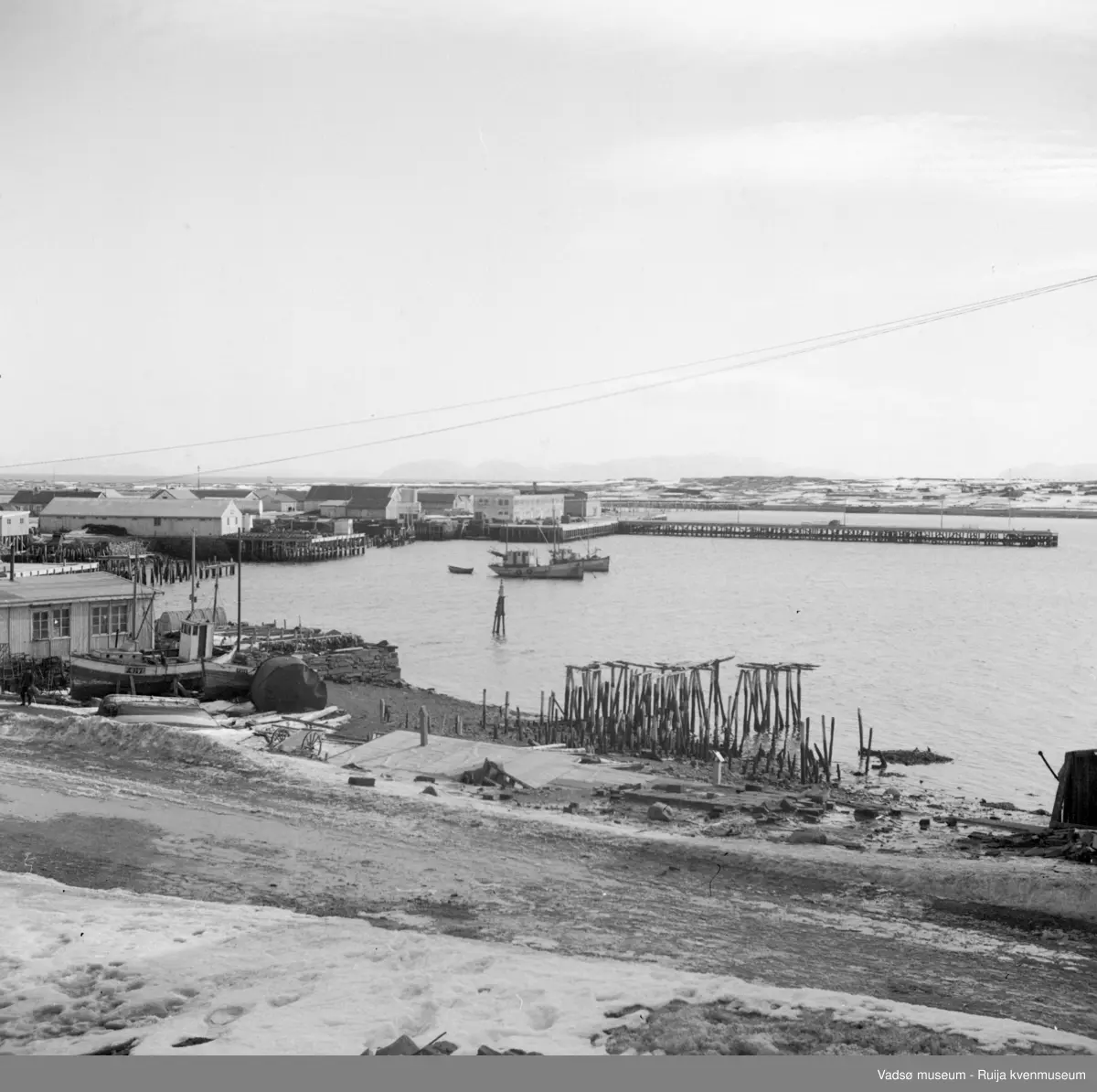 Vadsø. Bildet tatt mot sør med Vadsøya i bakgrunnen. Ser også fjellene på "sørsida". Nærmet sees rester etter kaianlegg og fiskebruk. En sjark ligger på land mens 2 sjarker er fortøyd i havna. Det er bygd nye kaier, lagerbygnigner og forretningsbygg nedover mot Ørtangen. På Aarsætherkaia er det hvite havnebygget og Aarsætherbruket kommet opp. Dampskipskaia ("hurtigrutekaia") stikker ut i sjøen.