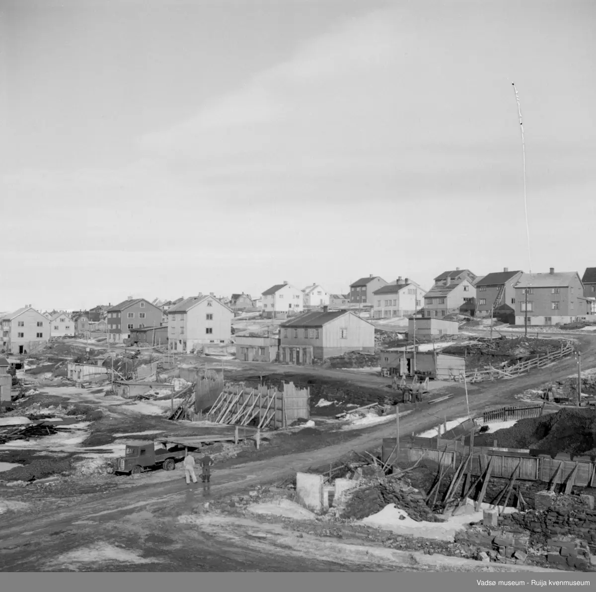 Vadsø sentrum 1951. Bildet tatt mot nordvest. 2 menn står i krysset Tollbugata/Kaigata. Gatene ovenfor er Havnegata og Nyborgveien. Vi ser en rekke bolighus med sokkeletg er gjenreist. Ser også brakker. Sentrum nærmest med nåværende torg og rådhus ligger fortsatt i ruiner. Et kullager nederst til høyre