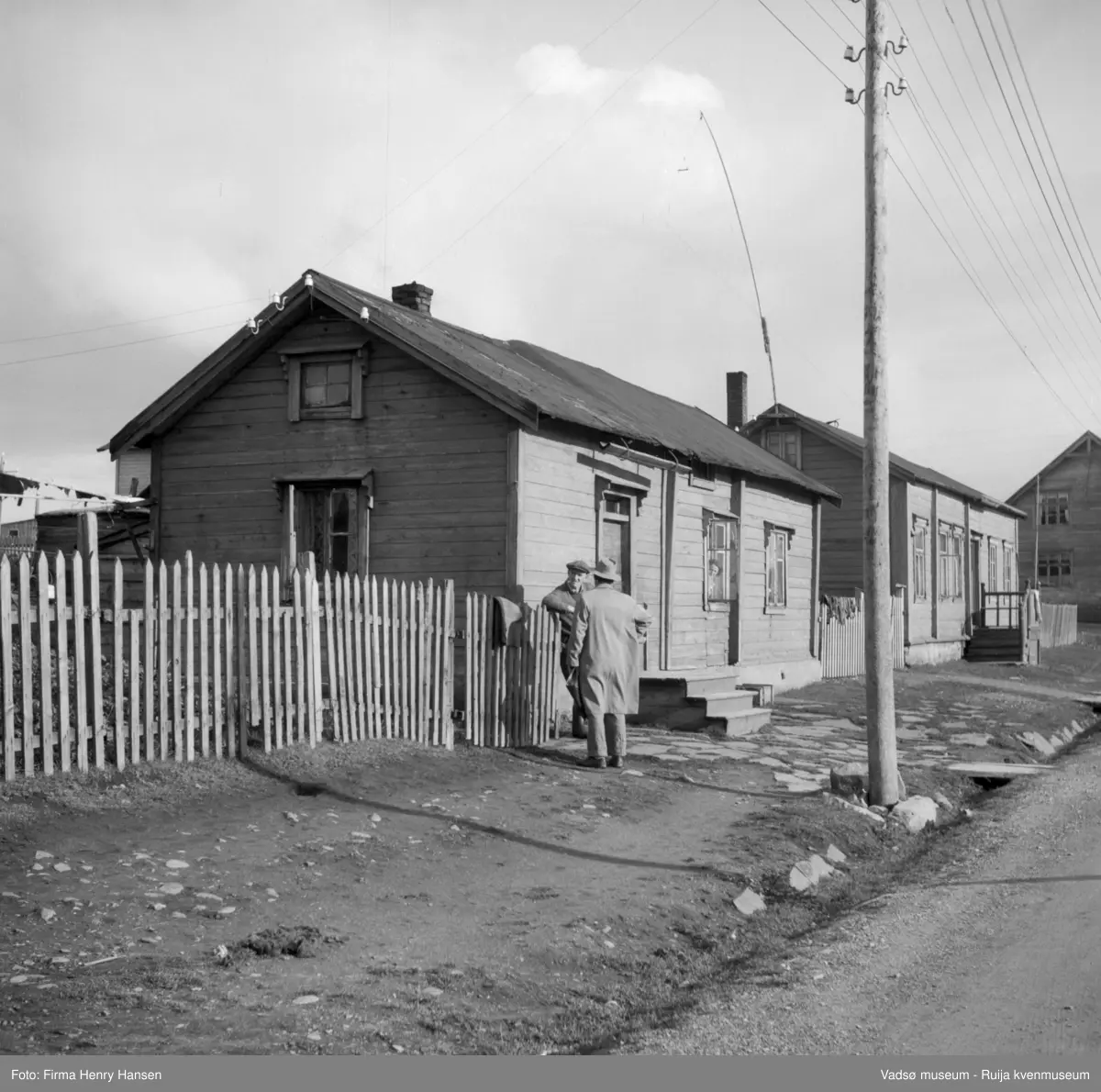 Vadsø, . Det nærmeste huset er Slettengata 25. Det ble bygd i 1845, ombygd fra ca 1948, det nye huset ble tatt  i bruk i 1949 og fikk et helt annet utseende, bl.a. ble nygygget på to etasjer. Husets eiere: Johanna og Felix Gallavara. Det gamle huset ble ikke revet, det ble bygd inn i det nye. På en av tømmerstokkene i stua som ble panelt over, skal det være risset inn årstallet 1711 og noen russiske bokstaver. Mann nr 1 fra venstre er Felix Gallavara (1905- ), kvinnen i vinduet er hans kone Johanna Gallavara (f.1907). Stolpen i forgrunnen er den samme som ble plassert der under krigen. Nabohuset til høyre er Slettengata 27.  Det ble bygd om i 1968 av nye eiere Irmgard og Jan Kåre Gallavara.
Kilde:  Trond Ballo: Vadsø bys eiendomshistorie. 2013.