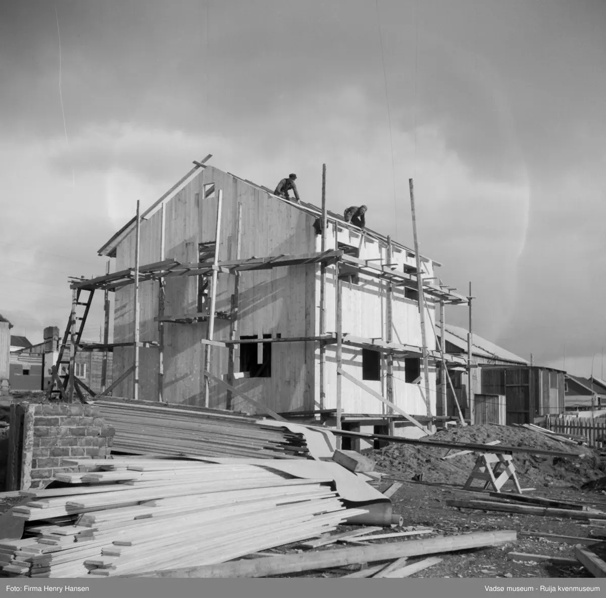 Vadsø. Bildet antas tatt mot vest. Gjenreisingshus i 2 etg under oppføring. 2 snekkere arbeider på taket. Det er satt opp stillas og byggematerialer ligger i forgrunen.