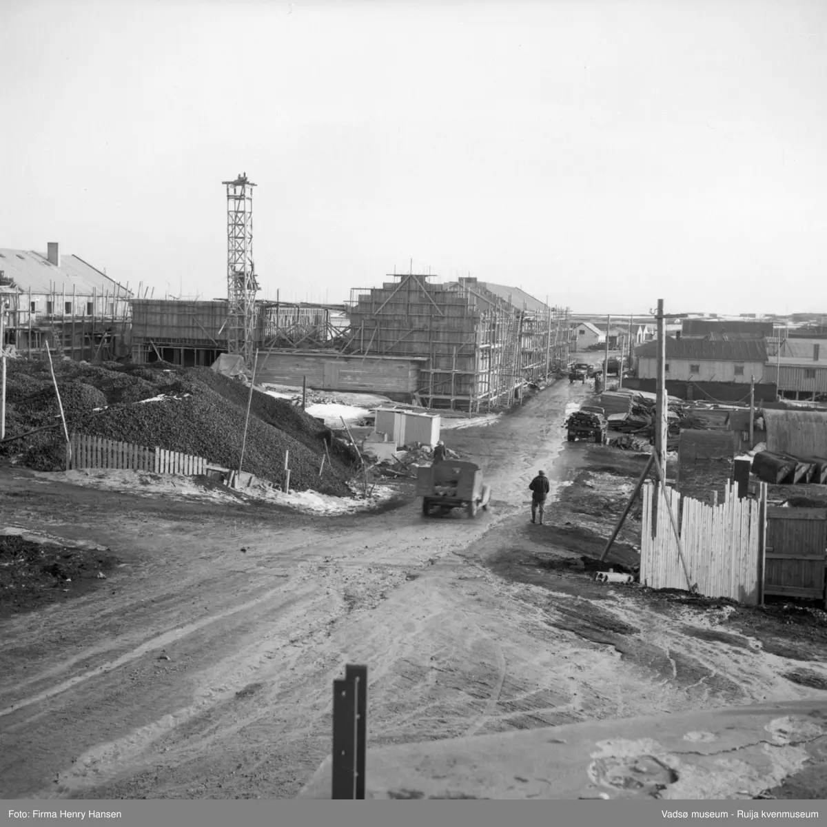 Vadsø sentrum. Gjenreisninga av Sentrum i full gang. Forretningsbyggene rundt Torget, øst for Tollbugata ; bla  Rolf Hansen, Henry Hansen og Baker Henriksen under oppføring. Bildet er tatt øverst i Tollbugata mot syd, og vi ser lastebiler med materialer kjører ivårløysninga.
