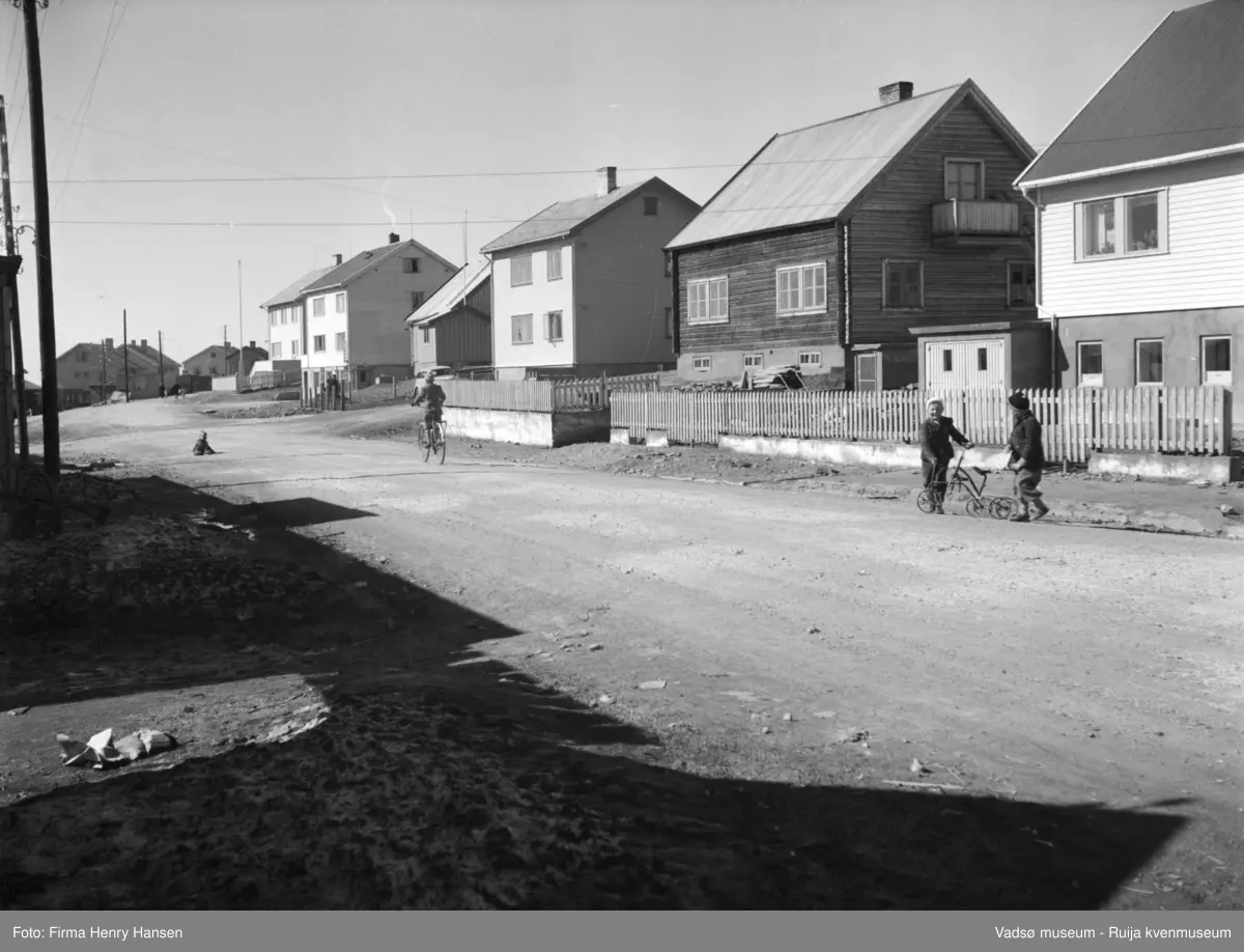Vadsø mai 1951. Oscarsgata vestover, fortsetter i Nyborgveien. Gjenreisningshus, både boliger og forretningsbygg, bl.a. Aksel Niskas bygg med kiosk i sokkeletasjen. Det nærmeste huset har lensmannskontoret i sokkeletasjen.