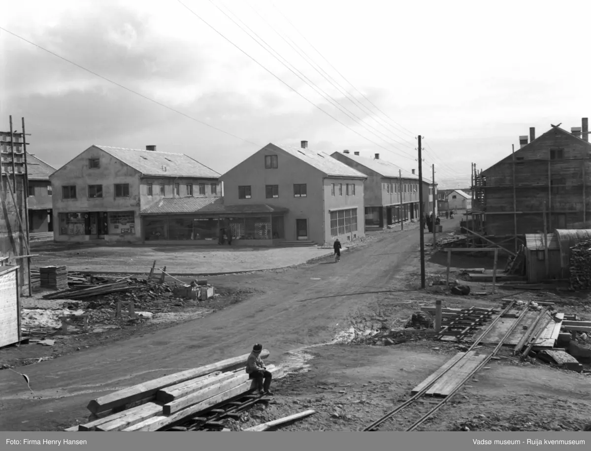 Vadsø mai 1951. Gjenreisninga i full gang i Sentrum. Bildet er tatt mot syd med Tollbugata midt i bildet. I venstre billedkant Vadsø Rådhus under oppføring. På Torget forretningsbyggene tilhørende Rolf Hansen og Henry Hansen. Lengre ned på østre side av Tollbugata H.F.Esbensens forretningsbygg. Vest for Tollbugata ses bl.a. forretningsbygg under oppføring.