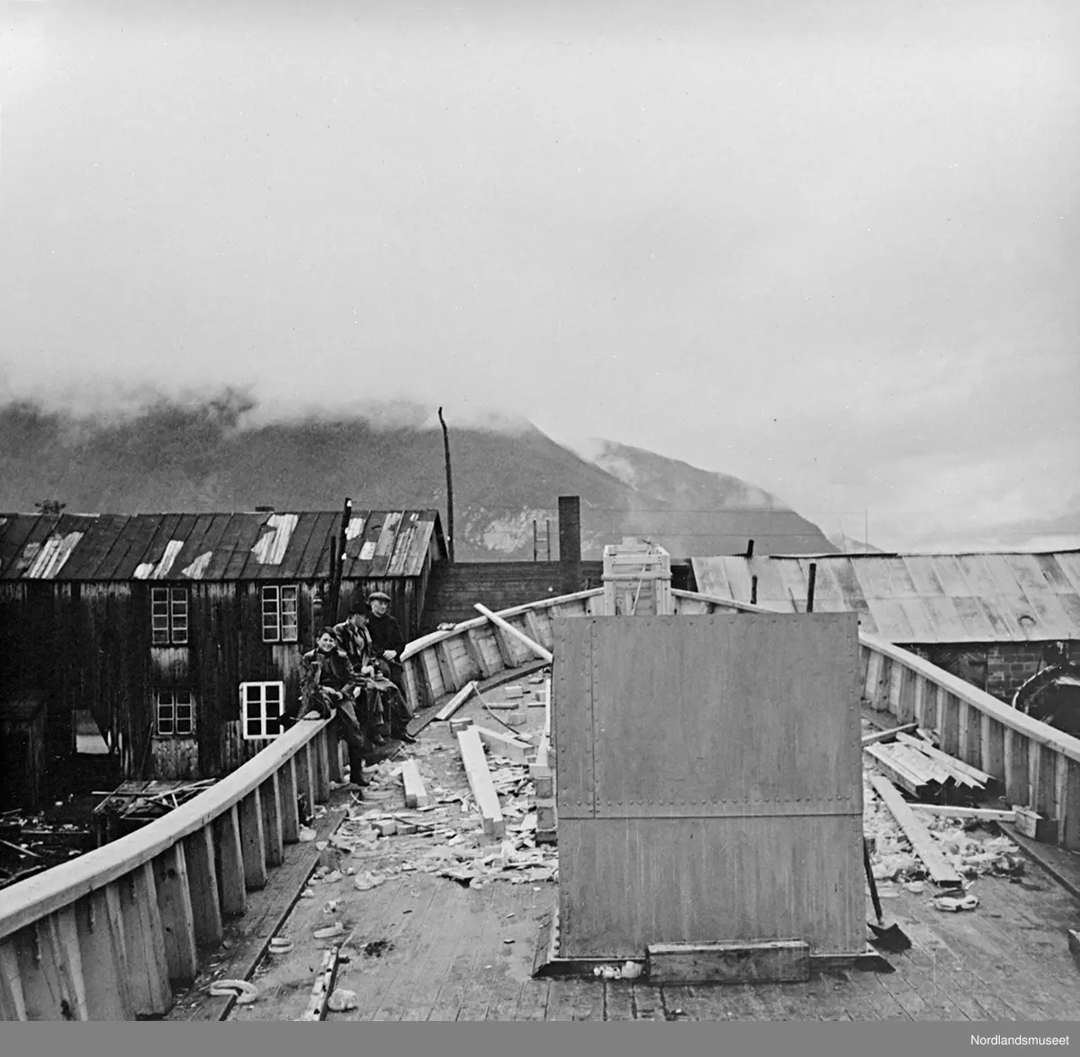 Slip, båtbyggeri. En trebåt under reparasjon/ombygging. Bildet er tatt ombord i båten mens den står på land. Bildet er tatt mot forstevnet, og viser dekksplanker, rekke og rekkstøtter. Tre personer sitter på rekklenningen. I bakgrunnen vises bygninger, sannsynligvis verkstedsbygg og administrasjon i tilknytning til slipen/båtbyggeriet.