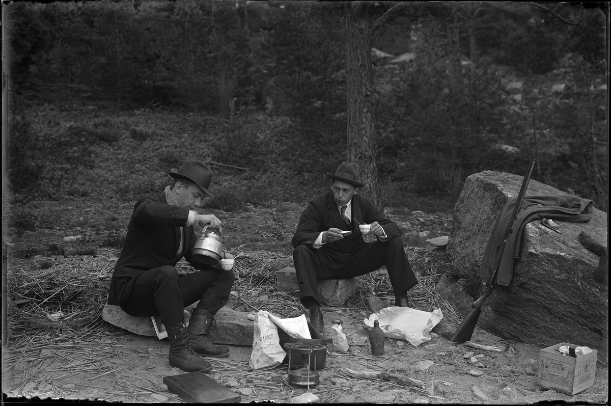 Hugo Larsson och Harald Olsson tar en fikapaus under en fiske och jakttur. På bilden sitter de på varsin sten med kaffekopp i handen. Harald häller upp kaffe ur en kanna och framför dem står ett Primuskök, en påse från A. Olssons specerier mm. Ett gevär står lutad mot en stor sten och på stenen ligger en rock, en pipa, tobak och en död ekorre.