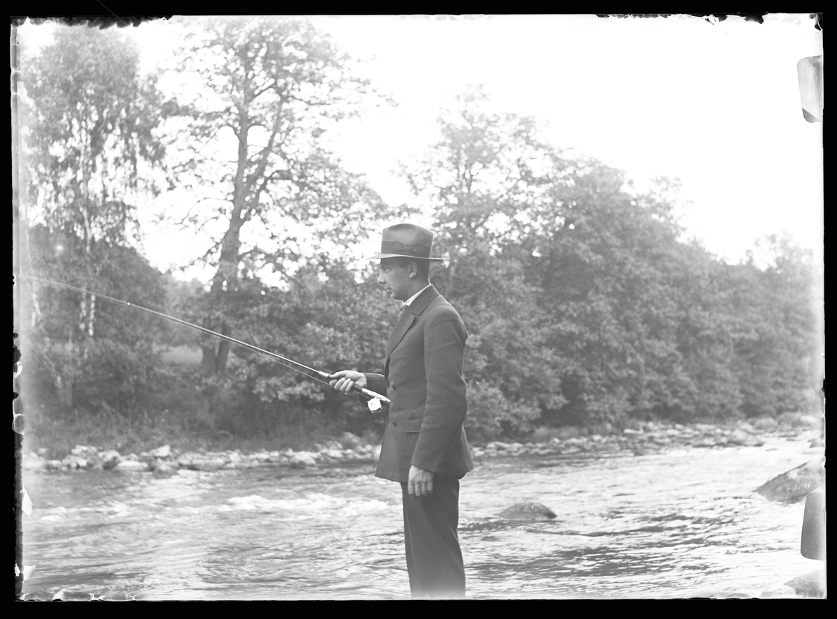 Harald Olsson klädd i hatt och kostym står vid vattnet och fiskar.