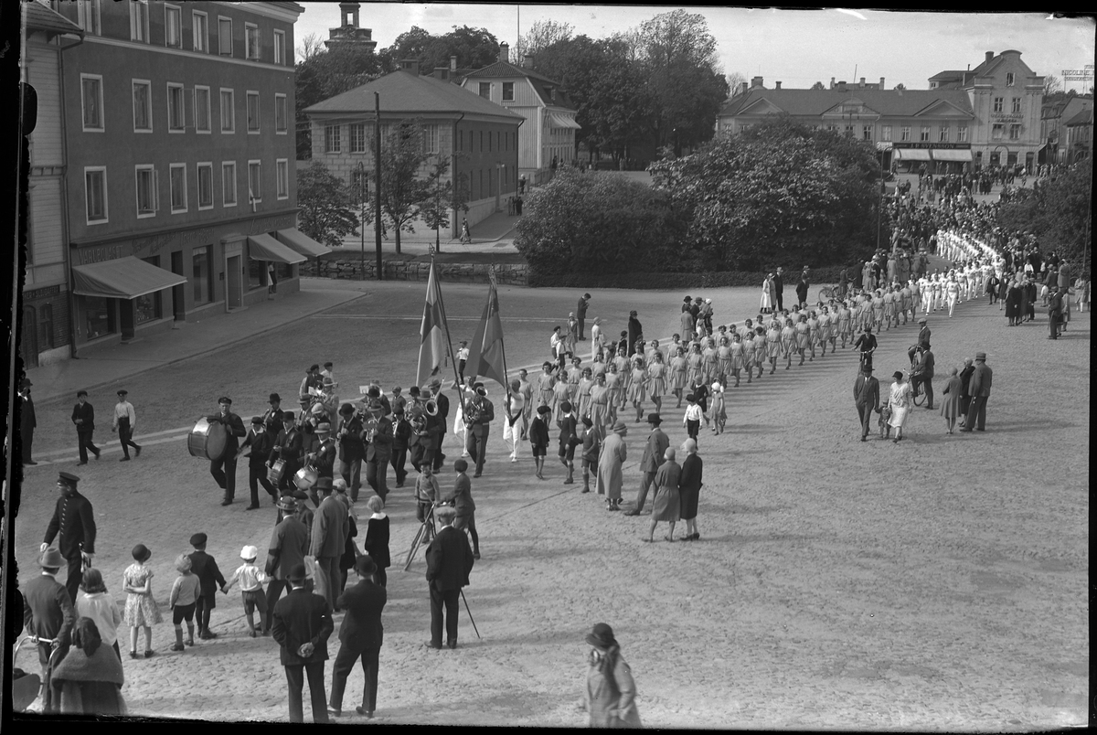 Musikanter och gymnaster går i en parad över Stora och Lilla torget. Runt om står människor och tittar på. Paraden hölls i samband med Gymnastikfesten på Idrottsplattsen.